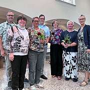 Eine Gruppe von sieben Personen posiert mit Blumen in einem hellen Raum.