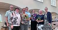 Eine Gruppe von sieben Personen posiert mit Blumen in einem hellen Raum.
